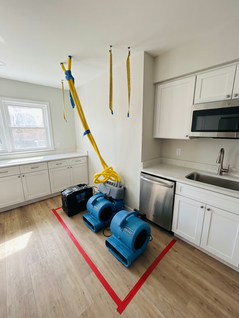 Air movers and a dehumidifier running inside a taped-off drying zone in a kitchen